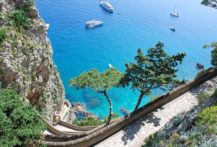 Caprin saari ja näkymä jyrkänteeltä kristallinkirkaaseen poukamaan jossa veneitä kauniina kesäpäivänä.