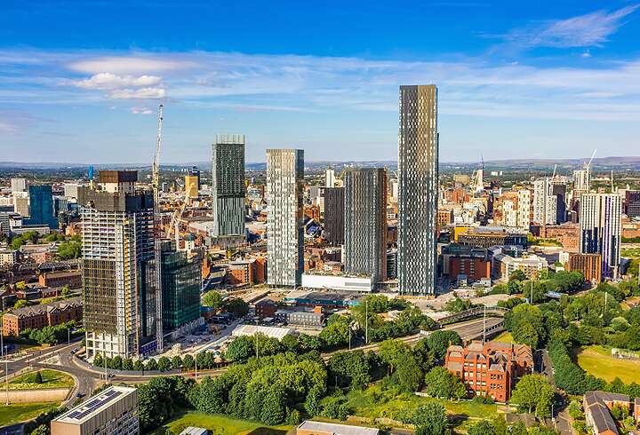Ilmakuva Manchesterin keskustasta, jossa näkyy moderneja pilvenpiirtäjiä, vehreitä puistoalueita ja kaupungin kaupunkirakennetta kirkkaana kesäpäivänä.