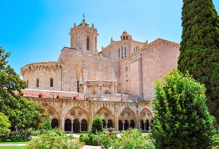 Espanjan Kataloniassa sijaitseva Tarragonan katedraali kylpee kirkkaassa kesäisessä auringonpaisteessa kauniiden sypressipuiden keskellä.